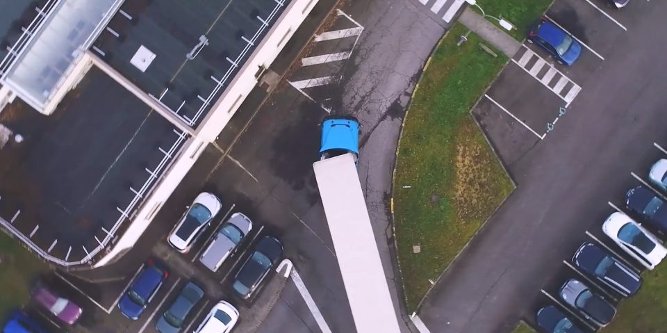 Vue plongeante sur un site industriel avec bâtiment, allée piétonne, parking et poubelles bleues.