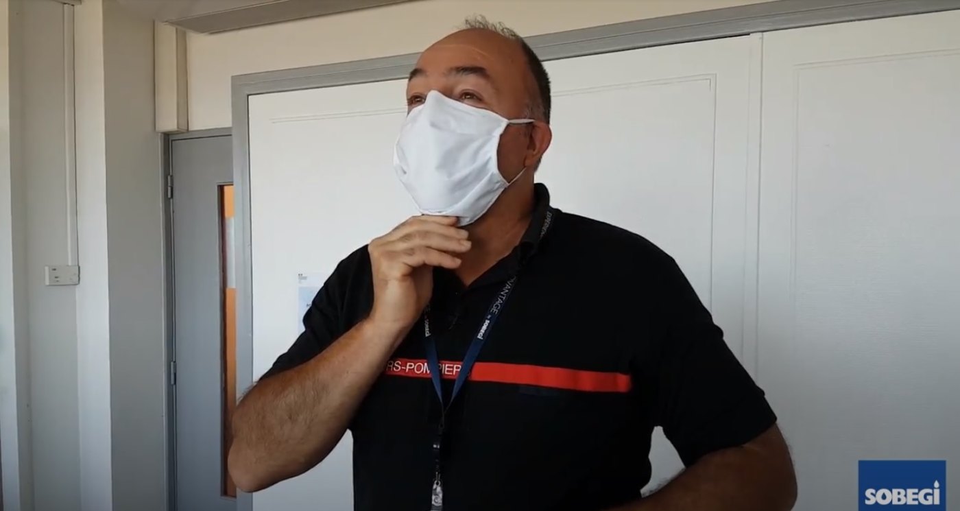 Homme portant un masque blanc et un lanyard, donnant des instructions devant une porte blanche.